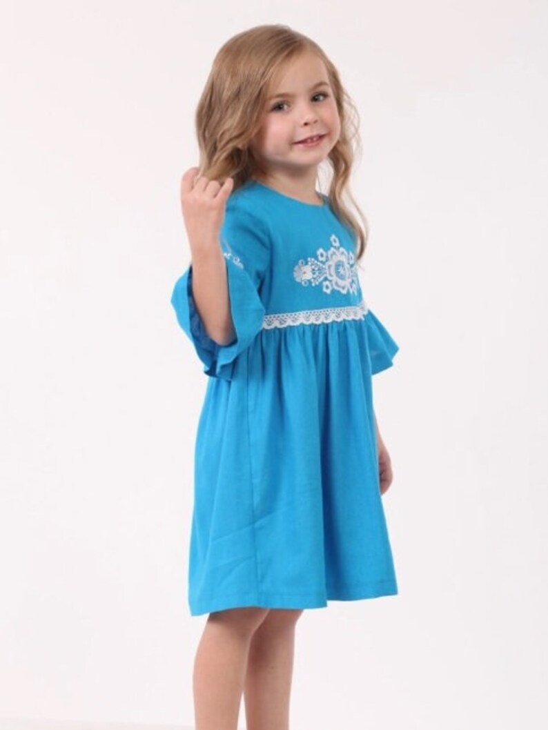 K&ouml;nnte beinhalten: Ein junges M&auml;dchen tr&auml;gt ein blaues Kleid mit wei&szlig;en Stickereien und einem wei&szlig;en Spitzenbesatz. Das Kleid hat einen R&uuml;schen-Saum und 3/4-&Auml;rmel.