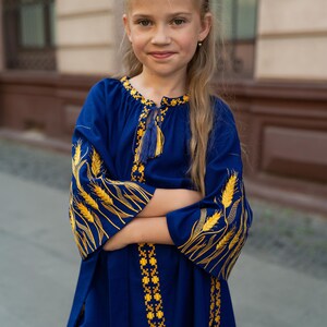 K&ouml;nnte beinhalten: Blaues Kleid mit gelber Stickerei. Das Kleid hat weizen&auml;hnliche Designs auf den &Auml;rmeln und dekorative Muster entlang des Halsausschnitts und des Saums. Das Kleid hat ein Quastendetail und wird von einer jungen Person getragen.