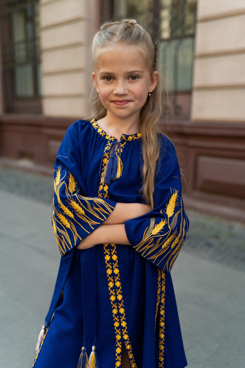 K&ouml;nnte beinhalten: Blaues Kleid mit gelber Stickerei. Das Kleid hat weizen&auml;hnliche Designs auf den &Auml;rmeln und dekorative Muster entlang des Halsausschnitts und des Saums. Das Kleid hat ein Quastendetail und wird von einer jungen Person getragen.