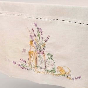 May include: Cream-colored linen tablecloth with embroidered lavender sprigs, bottles, and a lemon. The embroidery features purple, green, and yellow accents. The text "Lavender & Lemon" is embroidered on the tablecloth.
