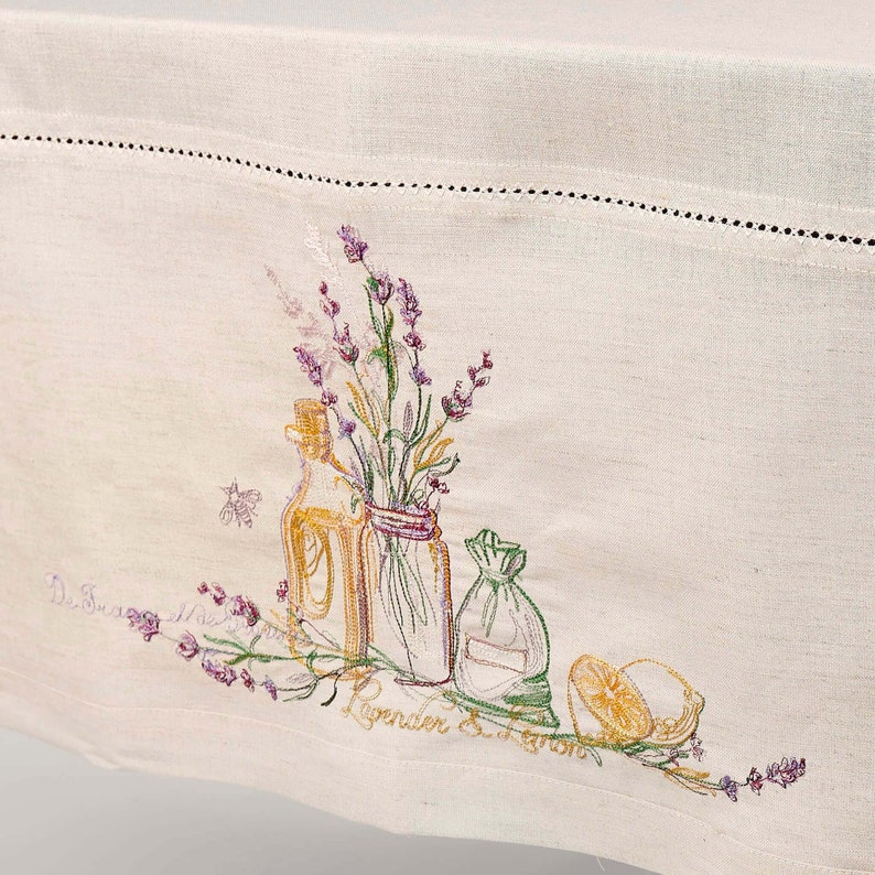 May include: Cream-colored linen tablecloth with embroidered lavender sprigs, bottles, and a lemon. The embroidery features purple, green, and yellow accents. The text "Lavender & Lemon" is embroidered on the tablecloth.