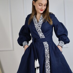 May include: A navy blue linen dress with white embroidery and tassel details. The dress has a V-neckline, long sleeves, and a wrap-style bodice. The dress is cinched at the waist with a matching belt.