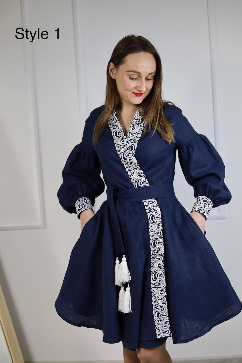 May include: A navy blue linen dress with white embroidery and tassel details. The dress has a V-neckline, long sleeves, and a wrap-style bodice. The dress is cinched at the waist with a matching belt.