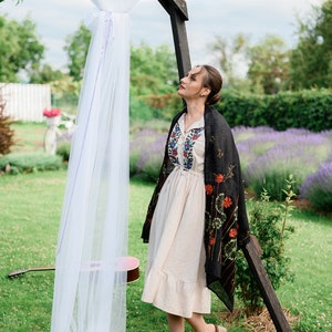 May include: A woman in a cream-colored dress with floral embroidery and a black shawl stands in a garden. A wooden arch is draped with white fabric. A guitar rests on the grass, and lavender flowers are in the background.