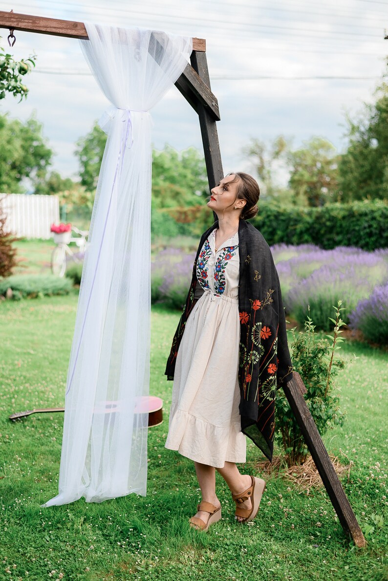 May include: A woman in a cream-colored dress with floral embroidery and a black shawl stands in a garden. A wooden arch is draped with white fabric. A guitar rests on the grass, and lavender flowers are in the background.