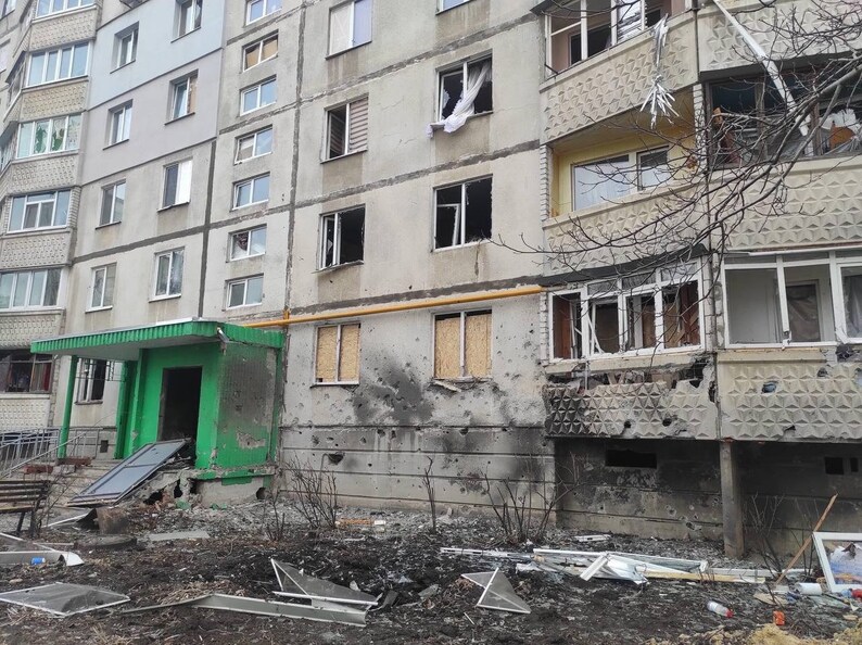 K&ouml;nnte beinhalten: Au&szlig;enansicht eines mehrst&ouml;ckigen Wohngeb&auml;udes mit erheblichen Sch&auml;den. Fenster sind zerbrochen, einige zugemauert. Die Fassade weist zahlreiche Einschussl&ouml;cher auf, und Tr&uuml;mmer liegen auf dem Boden. Der Eingang ist gr&uuml;n.
