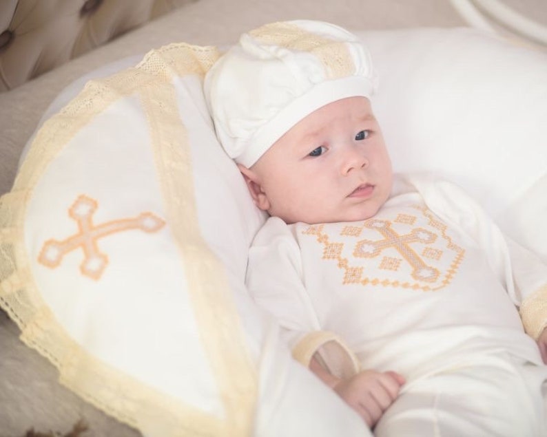 May include: A white baby christening gown with a gold embroidered cross on the front. The baby is wearing a white hat with a gold trim. The baby is lying on a white pillow with a gold lace trim.