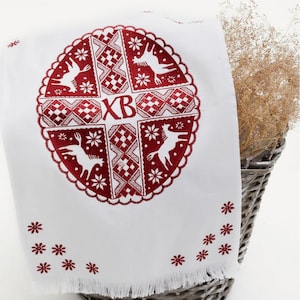 Op de afbeelding: Witte geborduurde keukendoek met een rood cirkelvormig ontwerp met een kruis met de letters "XB" in het midden. Het ontwerp bevat ook gestileerde dieren en geometrische patronen. De doek heeft een gefranst randje en is dubbelgevouwen.