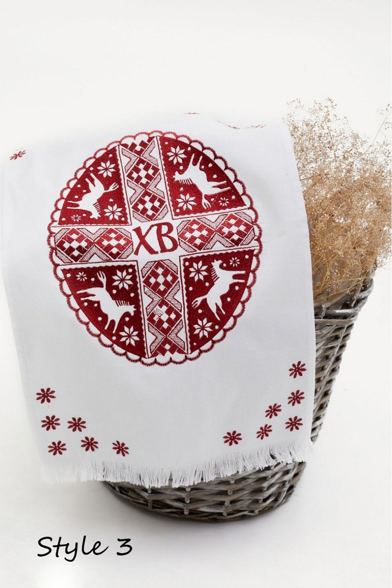 Op de afbeelding: Witte geborduurde keukendoek met een rood cirkelvormig ontwerp met een kruis met de letters "XB" in het midden. Het ontwerp bevat ook gestileerde dieren en geometrische patronen. De doek heeft een gefranst randje en is dubbelgevouwen.