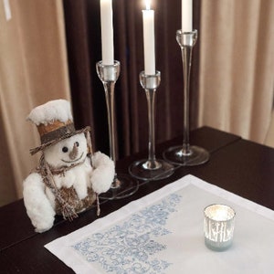 May include: A festive holiday scene featuring three lit white candles in elegant glass holders, a snowman figurine, and a small candle in a silver holder on a white tablecloth with a blue floral design. The scene is set on a dark wooden table.