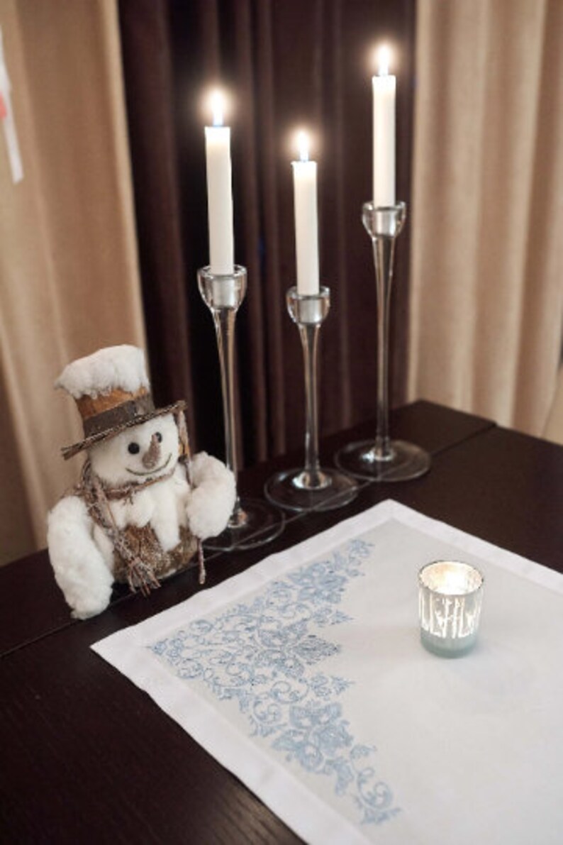May include: A festive holiday scene featuring three lit white candles in elegant glass holders, a snowman figurine, and a small candle in a silver holder on a white tablecloth with a blue floral design. The scene is set on a dark wooden table.