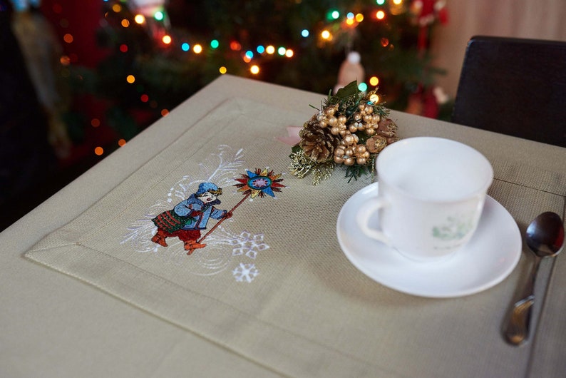 Op de afbeelding: Een beige placemat met een geborduurd volkskunstontwerp van een persoon die een stervormig object vasthoudt. Een witte kop en schotel, een lepel en een decoratief arrangement van gouden bessen en dennenappels liggen op de tafel.