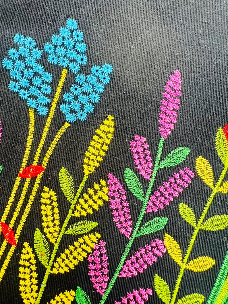 K&ouml;nnte beinhalten: Gestickte Blumenmuster auf schwarzem Stoffhintergrund. Das Design zeigt blaue, gelbe, gr&uuml;ne und rosa Blumen.