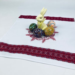 May include: White embroidered linen table runner with a red border. A white ceramic bunny sits on the runner surrounded by colorful Easter eggs.