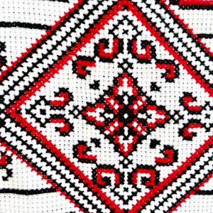 Puede incluir: Primer plano de una tela blanca con un detallado patr&oacute;n de punto de cruz. El dise&ntilde;o presenta formas geom&eacute;tricas en rojo y negro, creando un contraste llamativo. La costura es densa e intrincada, mostrando la artesan&iacute;a tradicional.