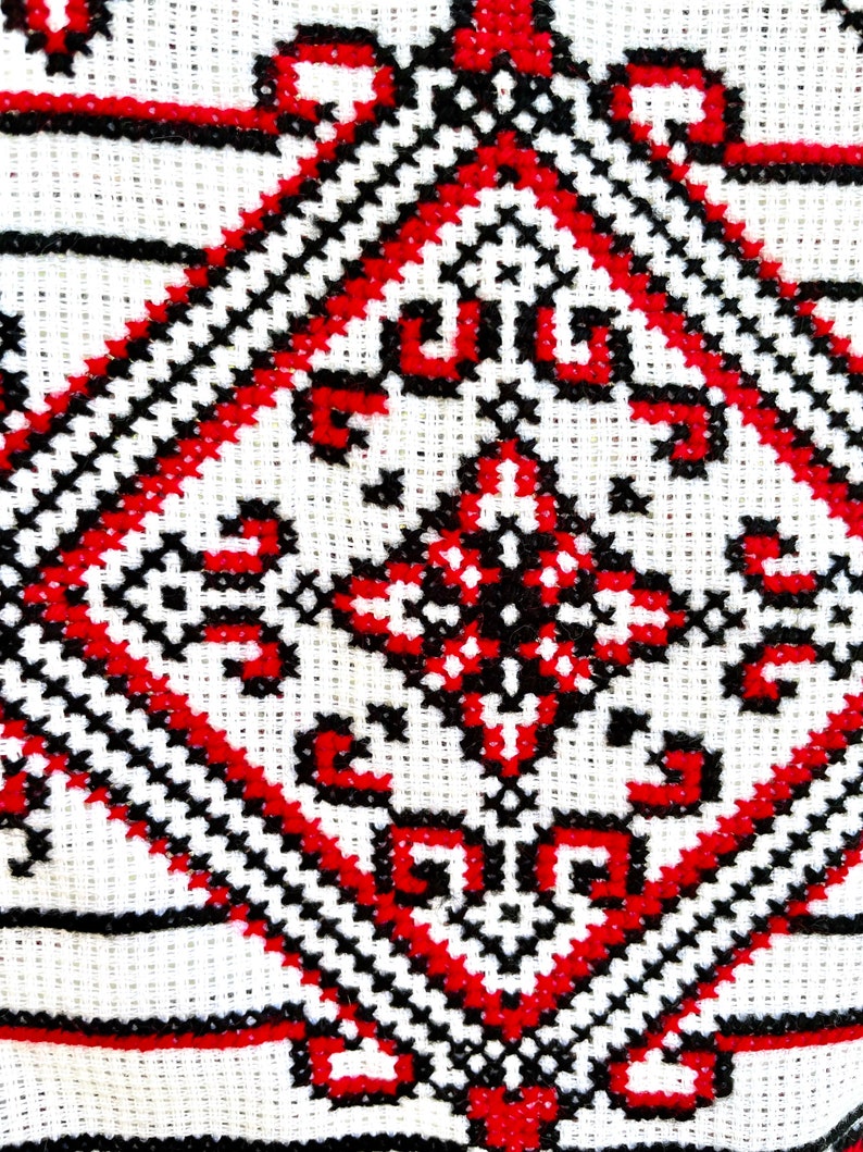 Puede incluir: Primer plano de una tela blanca con un detallado patr&oacute;n de punto de cruz. El dise&ntilde;o presenta formas geom&eacute;tricas en rojo y negro, creando un contraste llamativo. La costura es densa e intrincada, mostrando la artesan&iacute;a tradicional.