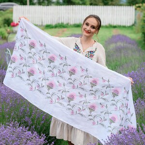 May include: A white scarf featuring a floral design of pink flowers and green stems. The scarf is displayed against a backdrop of purple flowers. The fabric appears to be lightweight.