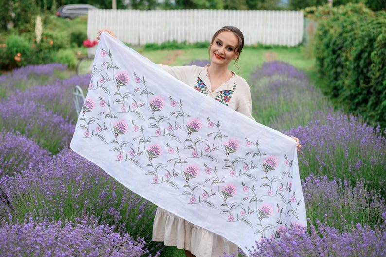 May include: A white scarf featuring a floral design of pink flowers and green stems. The scarf is displayed against a backdrop of purple flowers. The fabric appears to be lightweight.