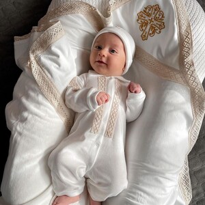 May include: A white baby outfit with lace trim and a white hat. The baby is lying on a white blanket with a gold cross embroidered on it.