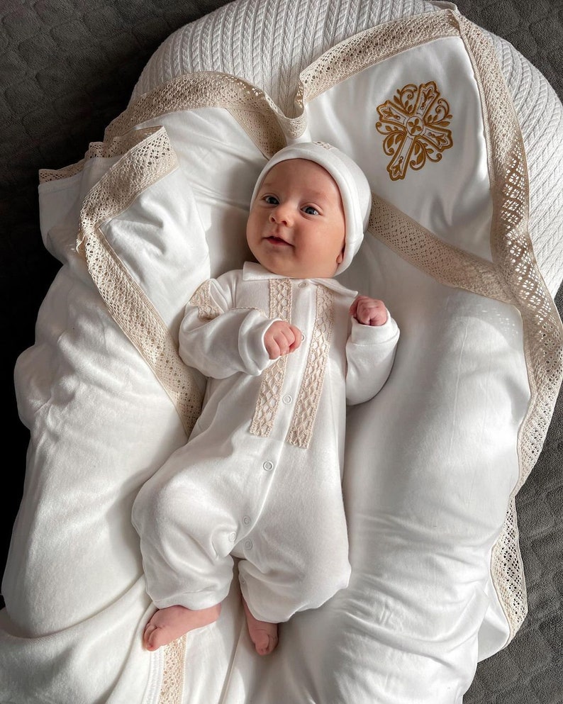 May include: A white baby outfit with lace trim and a white hat. The baby is lying on a white blanket with a gold cross embroidered on it.