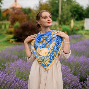 May include: A woman wearing a beige dress with a blue and yellow floral patterned scarf. She is standing in a field of lavender.