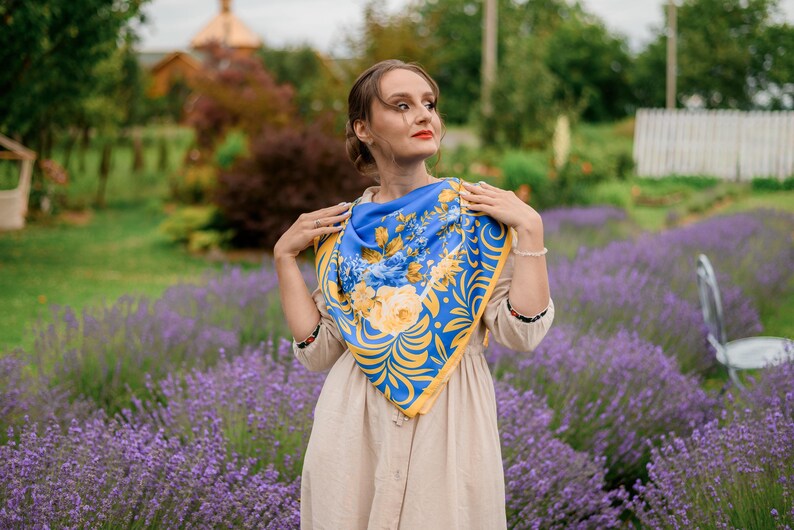May include: A woman wearing a beige dress with a blue and yellow floral patterned scarf. She is standing in a field of lavender.