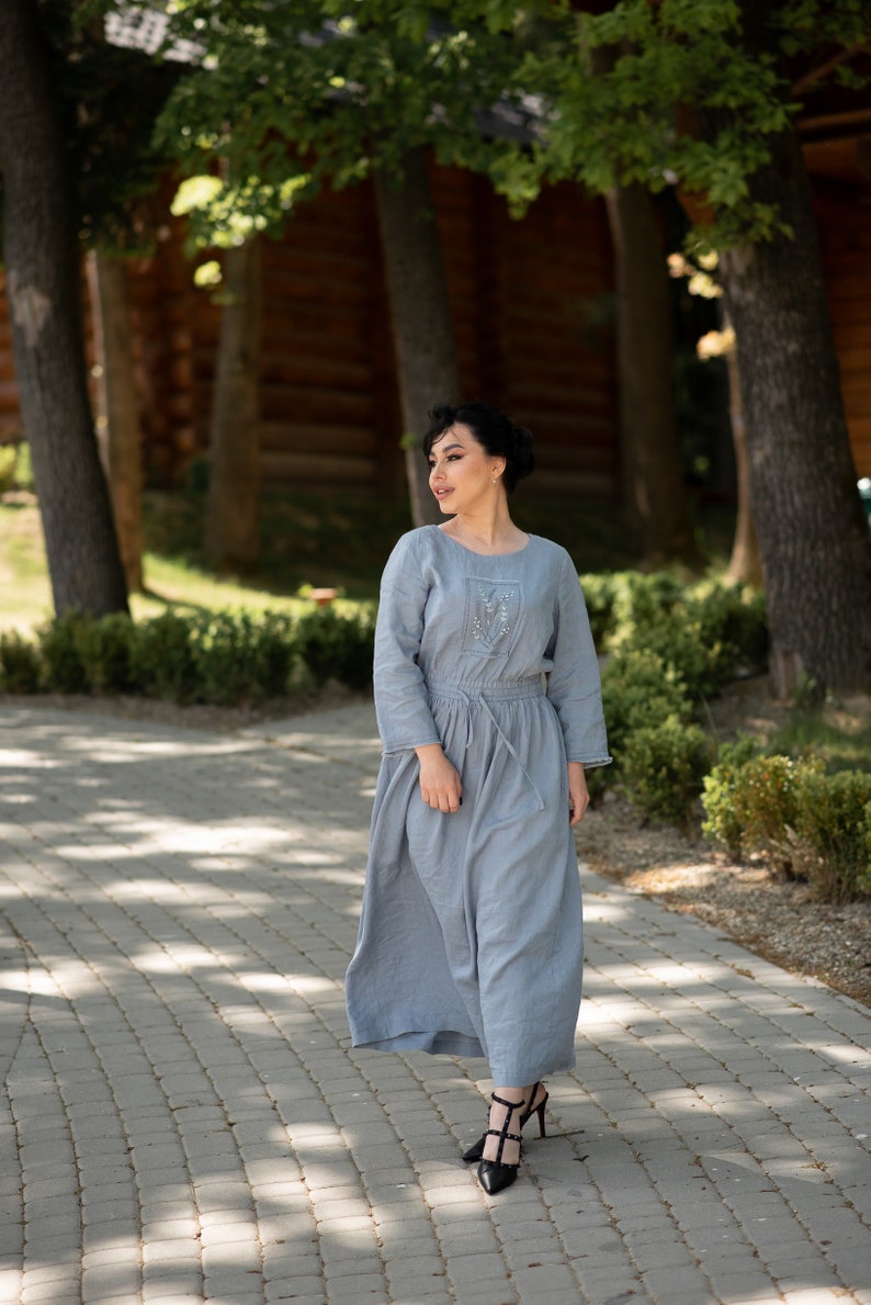 Może przedstawiać: Jasnoniebieska lniana sukienka z długimi rękawami i wiązaniem w talii. Sukienka ma delikatne hafty na gorsecie i jest zestawiona z czarnymi obcasami. Modelka stoi na brukowanej ścieżce, otoczona drzewami.