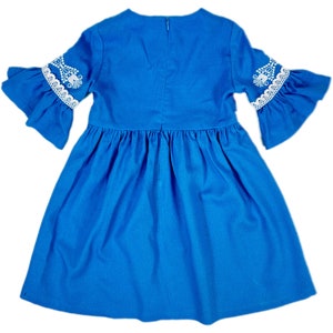 K&ouml;nnte beinhalten: Ein blaues Kleid mit wei&szlig;en Spitzenbesatz und Stickereien an den &Auml;rmeln. Das Kleid hat einen gerafften Rock und einen runden Ausschnitt.