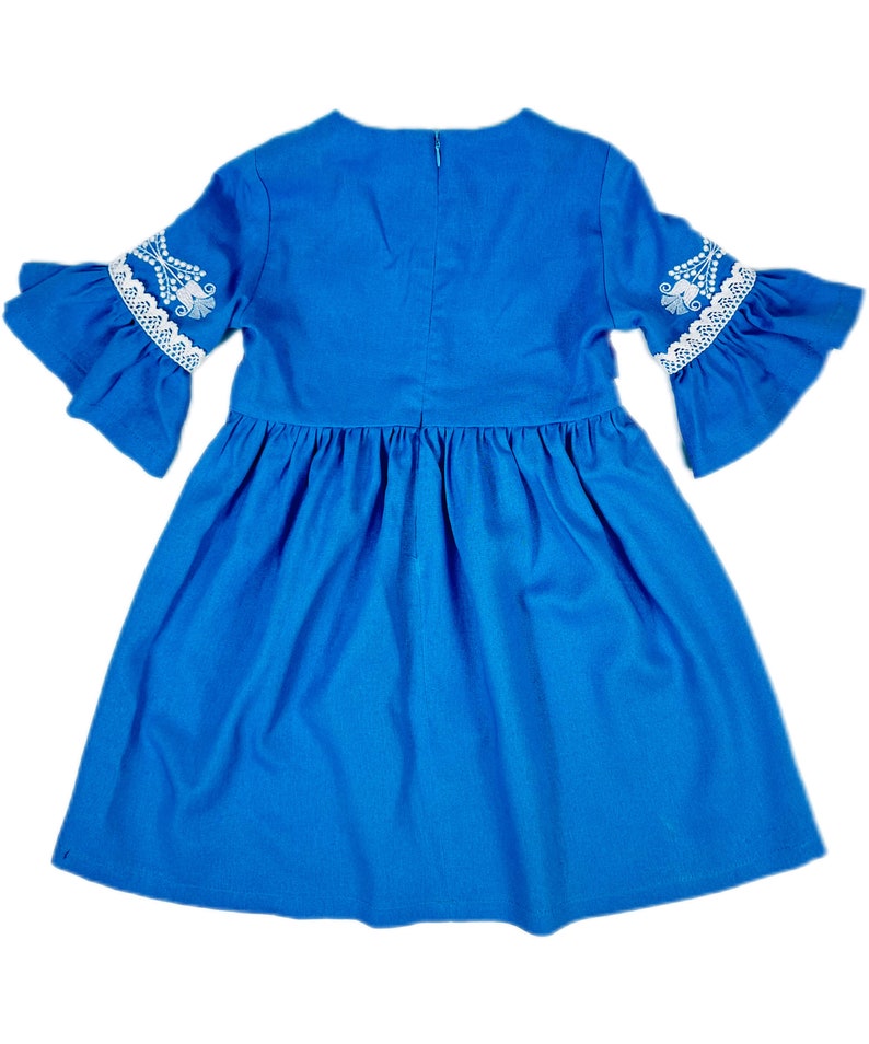 K&ouml;nnte beinhalten: Ein blaues Kleid mit wei&szlig;en Spitzenbesatz und Stickereien an den &Auml;rmeln. Das Kleid hat einen gerafften Rock und einen runden Ausschnitt.