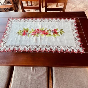 May include: A white table runner with a red and green floral embroidery design. The runner has a white lace border with red accents. The runner is on a dark brown wooden table.