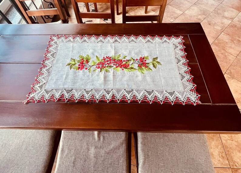 May include: A white table runner with a red and green floral embroidery design. The runner has a white lace border with red accents. The runner is on a dark brown wooden table.