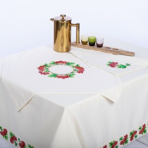 May include: A cream-colored tablecloth with embroidered red berries and green leaves. A gold-colored French press, a wooden serving tray with three small glasses, and a matching napkin are on the table. The tablecloth has a berry border.