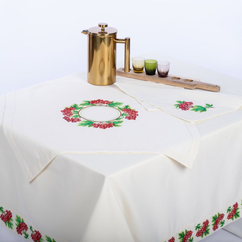 May include: A cream-colored tablecloth with embroidered red berries and green leaves. A gold-colored French press, a wooden serving tray with three small glasses, and a matching napkin are on the table. The tablecloth has a berry border.