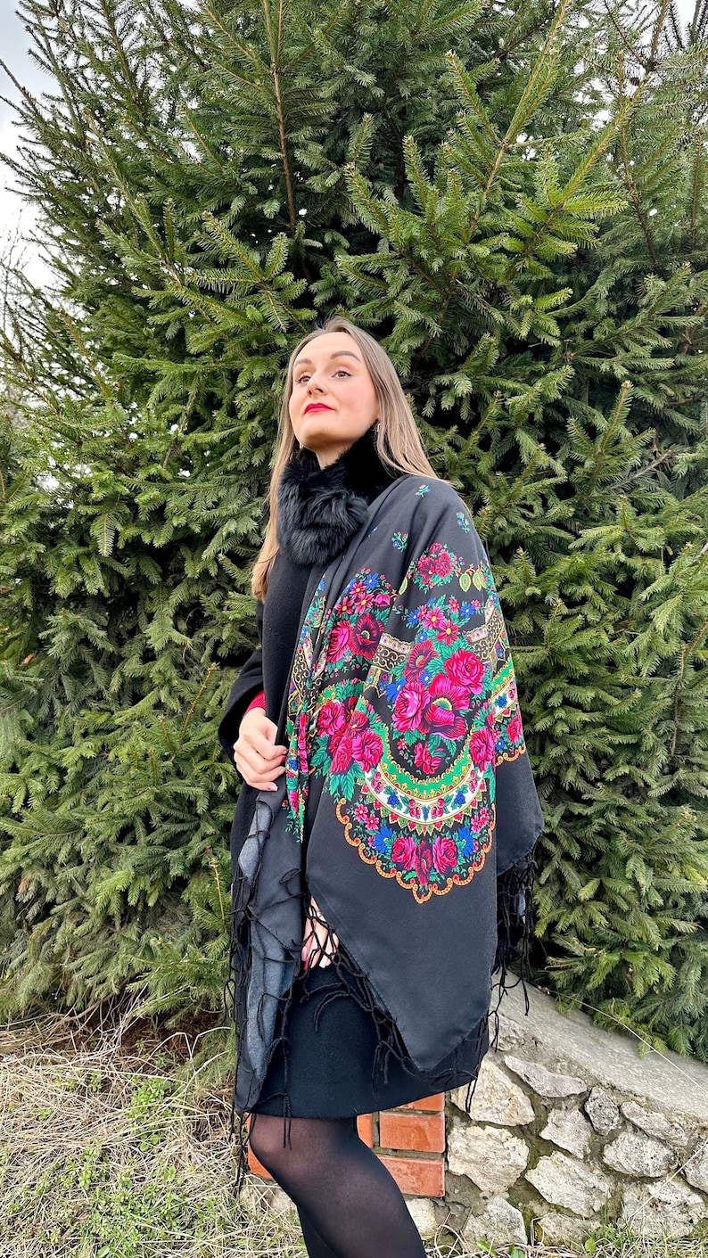 Puede incluir: Una mujer con un vestido negro y medias negras con un chal negro con un estampado floral colorido. El chal tiene flecos alrededor de los bordes. Est&aacute; de pie frente a un &aacute;rbol de hoja perenne verde.