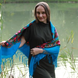 May include: A woman wearing a blue shawl with a floral pattern. The shawl has a fringe along the edges. The background is a lake with reeds.