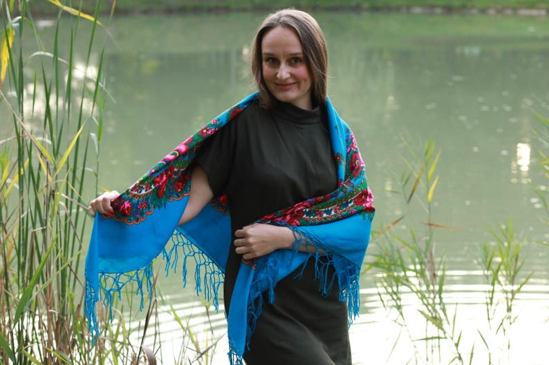May include: A woman wearing a blue shawl with a floral pattern. The shawl has a fringe along the edges. The background is a lake with reeds.