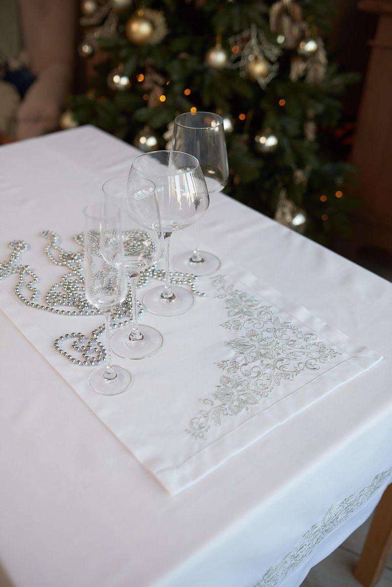 May include: A white tablecloth with a decorative embroidered runner and several wine glasses. The runner features an ornate silver floral design. Silver beaded garland is draped across the table, creating a festive holiday setting.