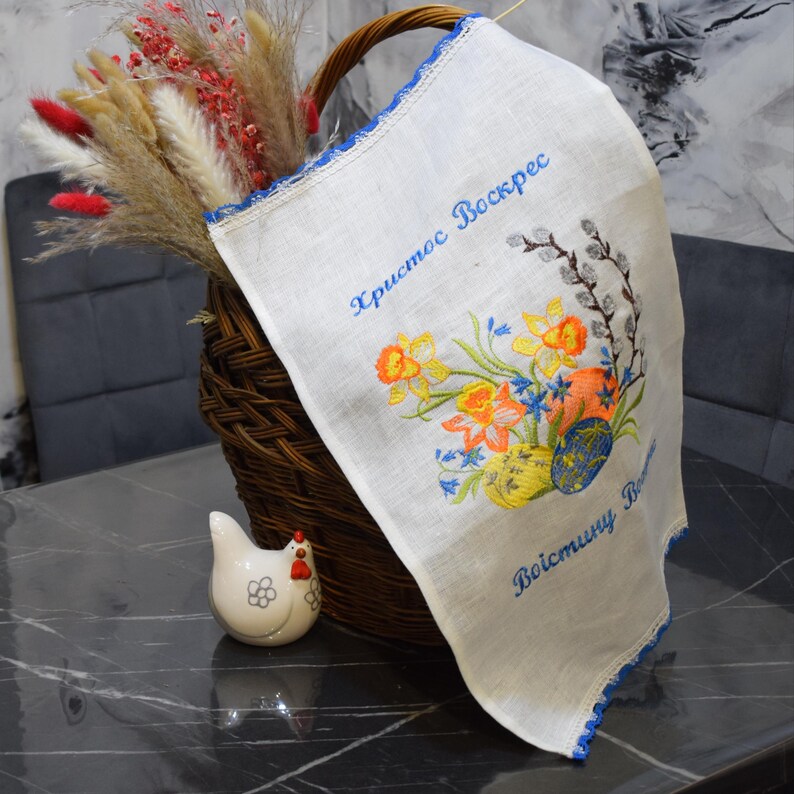 Puede incluir: Una toalla de t&eacute; de lino blanco con un borde bordado azul y un dise&ntilde;o floral. El dise&ntilde;o incluye narcisos, campanillas azules y huevos de Pascua. El texto "Христос Воскрес" (Cristo ha resucitado) est&aacute; bordado en hilo azul. La toalla est&aacute; doblada y colocada en una cesta de mimbre.