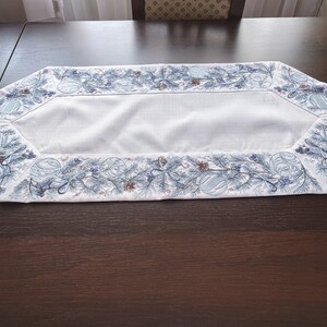 以下が含まれることがあります： 青と白の刺繍の縁取りが施された白いテーブルランナー。冬らしい花のデザインが施されています。縁取りには、松の枝、ベリー、オーナメントが繰り返しパターンで描かれています。