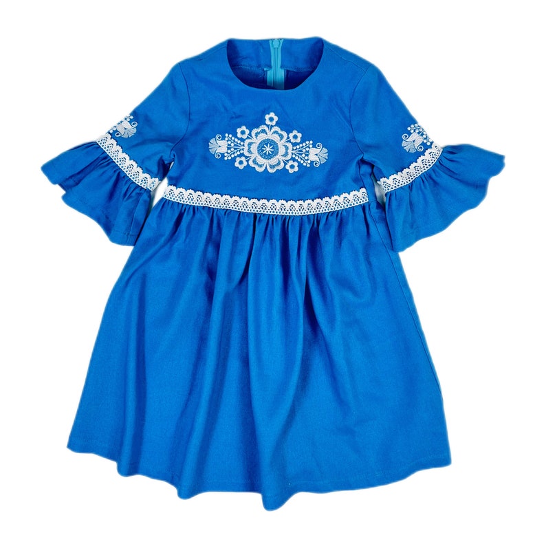 K&ouml;nnte beinhalten: Ein blaues Kleid mit wei&szlig;en Stickereien und Spitzenbesatz. Das Kleid hat einen runden Ausschnitt und 3/4 &Auml;rmel.