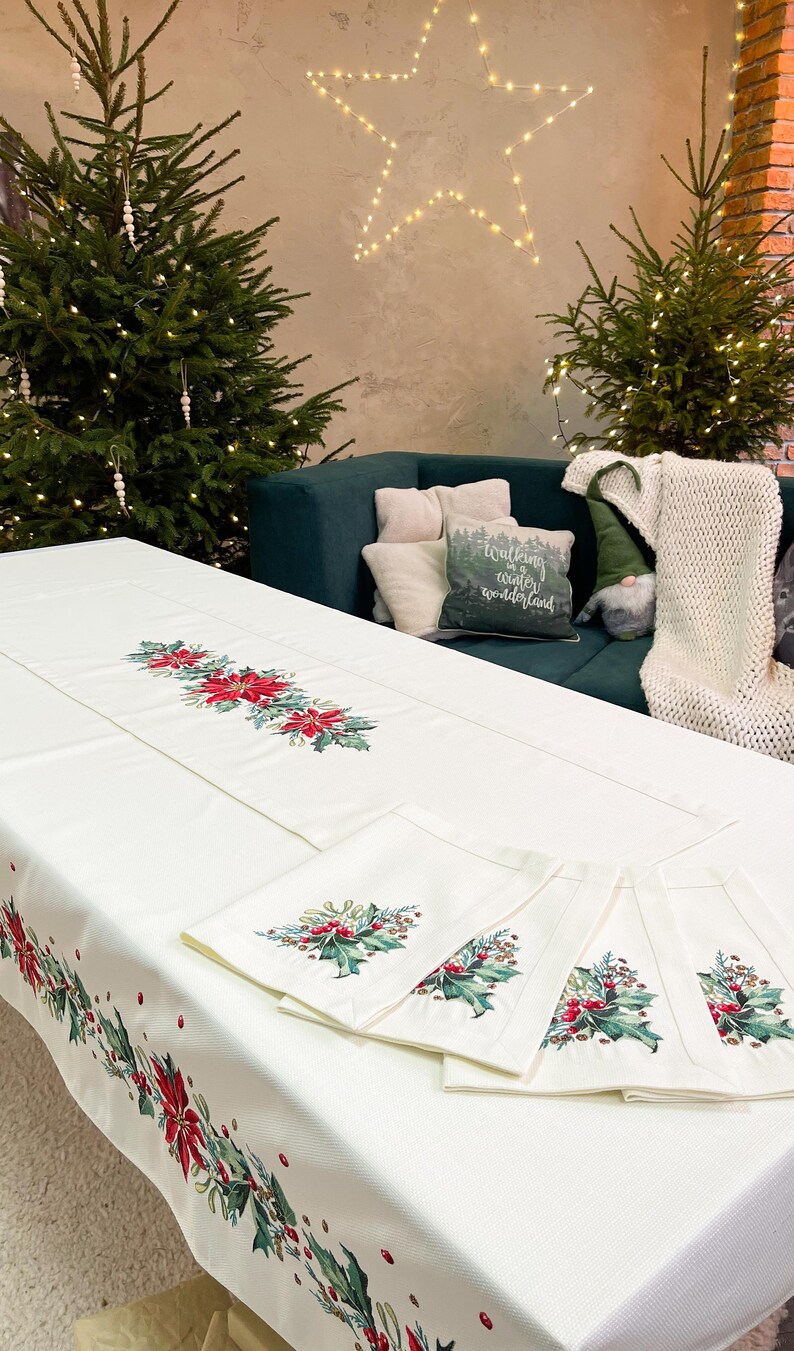 May include: White tablecloth with a red and green holly and poinsettia design. The tablecloth is set with matching napkins and a runner. The text "Walking in a winter wonderland" is on a cushion in the background.