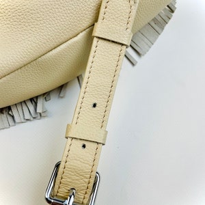 Puede incluir: Primer plano de un bolso de cuero color crema con un borde con flecos. El bolso presenta una correa de cuero a juego con una hebilla plateada. El cuero tiene una apariencia texturizada y la costura es visible.