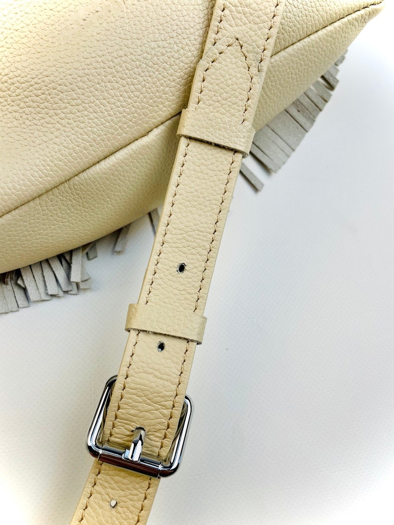 Puede incluir: Primer plano de un bolso de cuero color crema con un borde con flecos. El bolso presenta una correa de cuero a juego con una hebilla plateada. El cuero tiene una apariencia texturizada y la costura es visible.