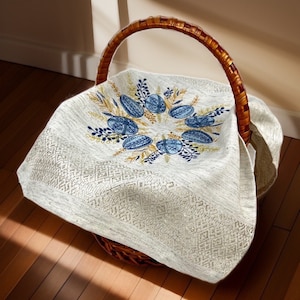 Puede incluir: Una cesta de mimbre tejida con un asa alta, forrada con un pa&ntilde;o color crema. El pa&ntilde;o est&aacute; bordado con dise&ntilde;os de huevos azules y dorados y detalles florales. La cesta est&aacute; sobre una superficie de madera.