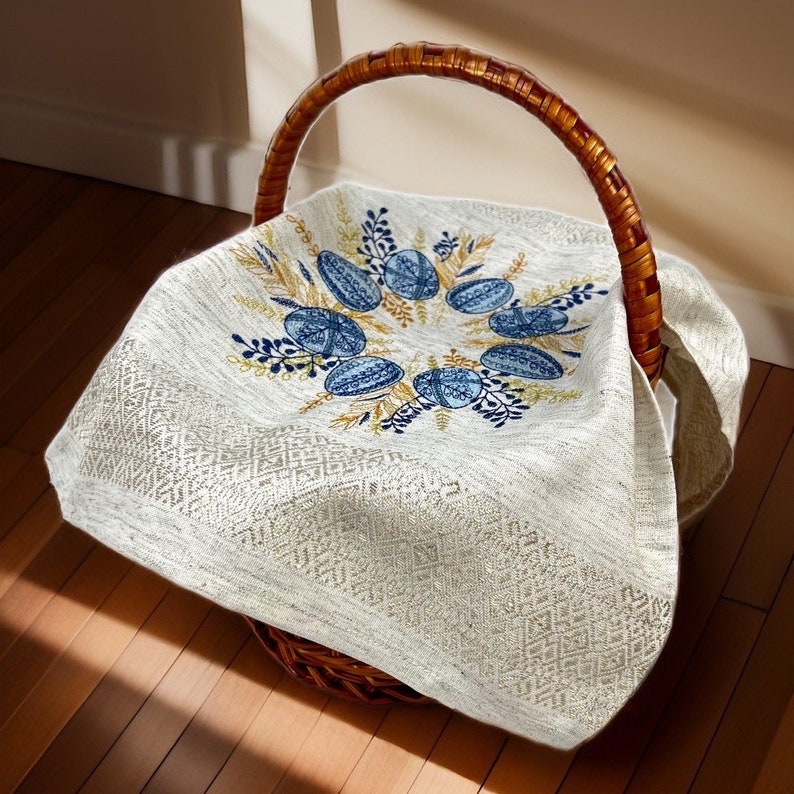 Puede incluir: Una cesta de mimbre tejida con un asa alta, forrada con un pa&ntilde;o color crema. El pa&ntilde;o est&aacute; bordado con dise&ntilde;os de huevos azules y dorados y detalles florales. La cesta est&aacute; sobre una superficie de madera.