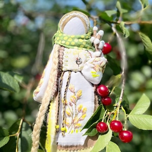 Puede incluir: Una mu&ntilde;eca de tela hecha a mano con cabello largo rubio, vistiendo un vestido blanco con bordados amarillos, una bufanda verde con lunares blancos y un cintur&oacute;n marr&oacute;n. La mu&ntilde;eca sostiene una rama con bolas de algod&oacute;n blancas y est&aacute; de pie frente a un cerezo con cerezas rojas.