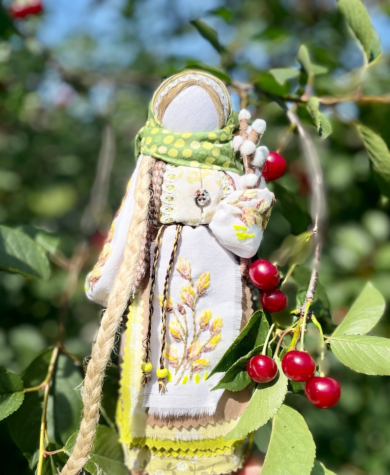 Puede incluir: Una mu&ntilde;eca de tela hecha a mano con cabello largo rubio, vistiendo un vestido blanco con bordados amarillos, una bufanda verde con lunares blancos y un cintur&oacute;n marr&oacute;n. La mu&ntilde;eca sostiene una rama con bolas de algod&oacute;n blancas y est&aacute; de pie frente a un cerezo con cerezas rojas.