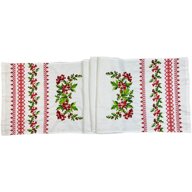 K&ouml;nnte beinhalten: Wei&szlig;er, bestickter Tischl&auml;ufer mit roten Beeren, gr&uuml;nen Bl&auml;ttern und braunen Eicheln. Der L&auml;ufer hat einen roten geometrischen Bord&uuml;rendesign. Die Stickerei ist auf dem wei&szlig;en Stoff zentriert.