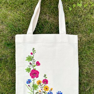 以下が含まれることがあります： 白いキャンバス地のトートバッグに、花柄の刺繍が施されています。刺繍には、ピンク、イエロー、ブルーの花と緑の葉が描かれています。