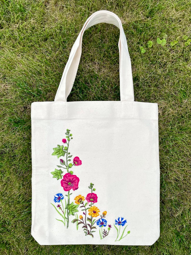 以下が含まれることがあります： 白いキャンバス地のトートバッグに、花柄の刺繍が施されています。刺繍には、ピンク、イエロー、ブルーの花と緑の葉が描かれています。